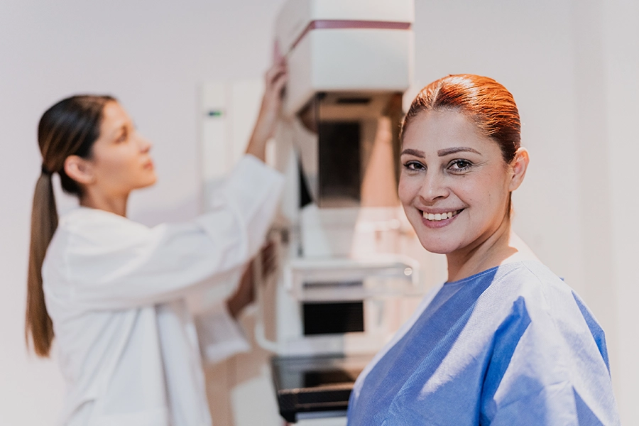 Patient undergoing a mammogram while a healthcare professional prepares the mammography machine for breast cancer screening.
